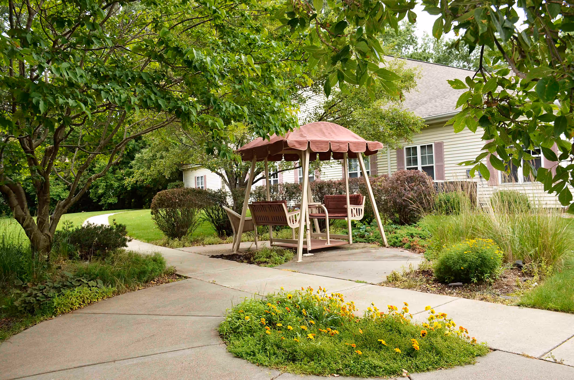 A peaceful outdoor garden area at Boden Senior Living Coon Rapids featuring a covered swing bench on a concrete patio surrounded by green trees, bushes, and flowering plants with a residential building in the background.