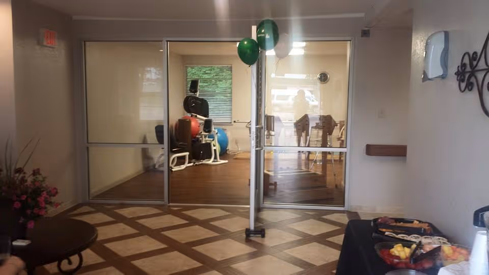 Interior lobby area with glass double doors opening to a room with exercise equipment and a table of refreshments nearby.