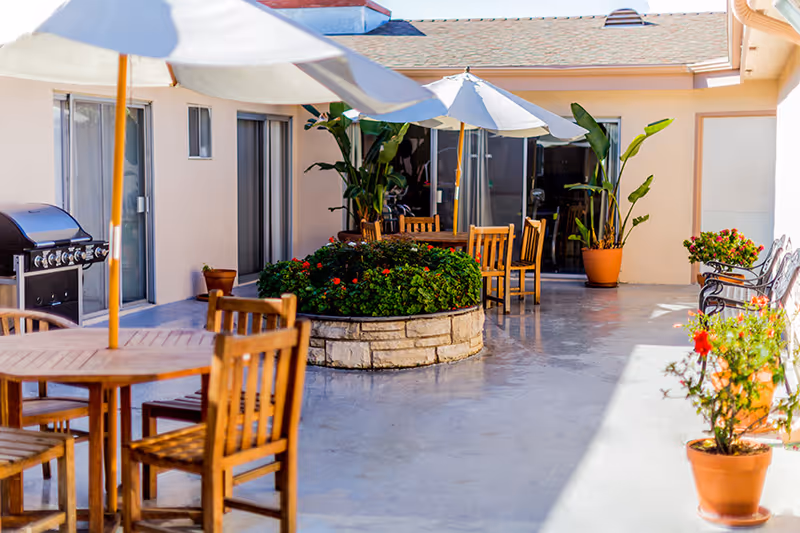 Outdoor patio area with wooden tables and chairs under large white umbrellas. There is a stone planter filled with green plants and red flowers in the center. Potted plants are placed along the edges near the building walls, and a barbecue grill is visible on the left side near sliding glass doors.