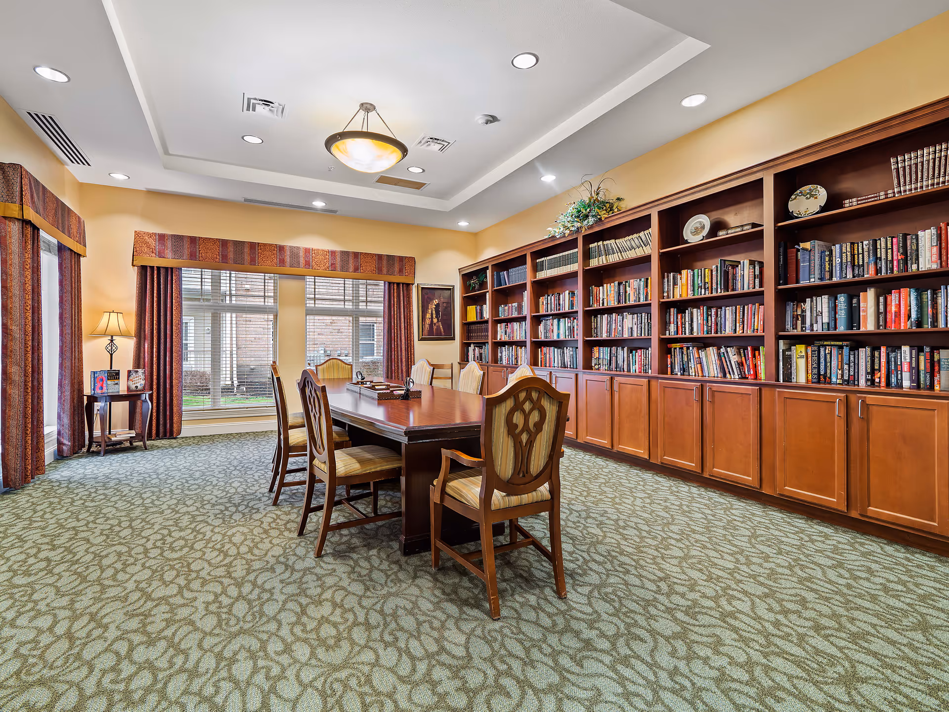 A well-lit library room with a long wooden table surrounded by six upholstered chairs. The room features a large built-in wooden bookshelf filled with books and decorative plates. There are large windows with patterned curtains allowing natural light to enter, and a small side table with a lamp and books. The ceiling has recessed lighting and a central hanging light fixture.