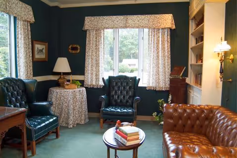 A cozy living room with dark green walls and a large window with floral curtains. The room features two dark green leather armchairs, a brown leather tufted sofa, a round table with a floral tablecloth and a lamp, a small round coffee table with books, and built-in bookshelves with a wall-mounted lamp.
