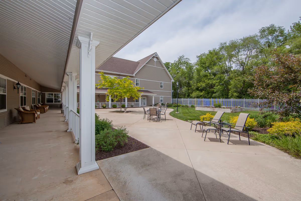 Covered walkway and outdoor courtyard with patio chairs, tables, and landscaping beside a multi-story senior living building.