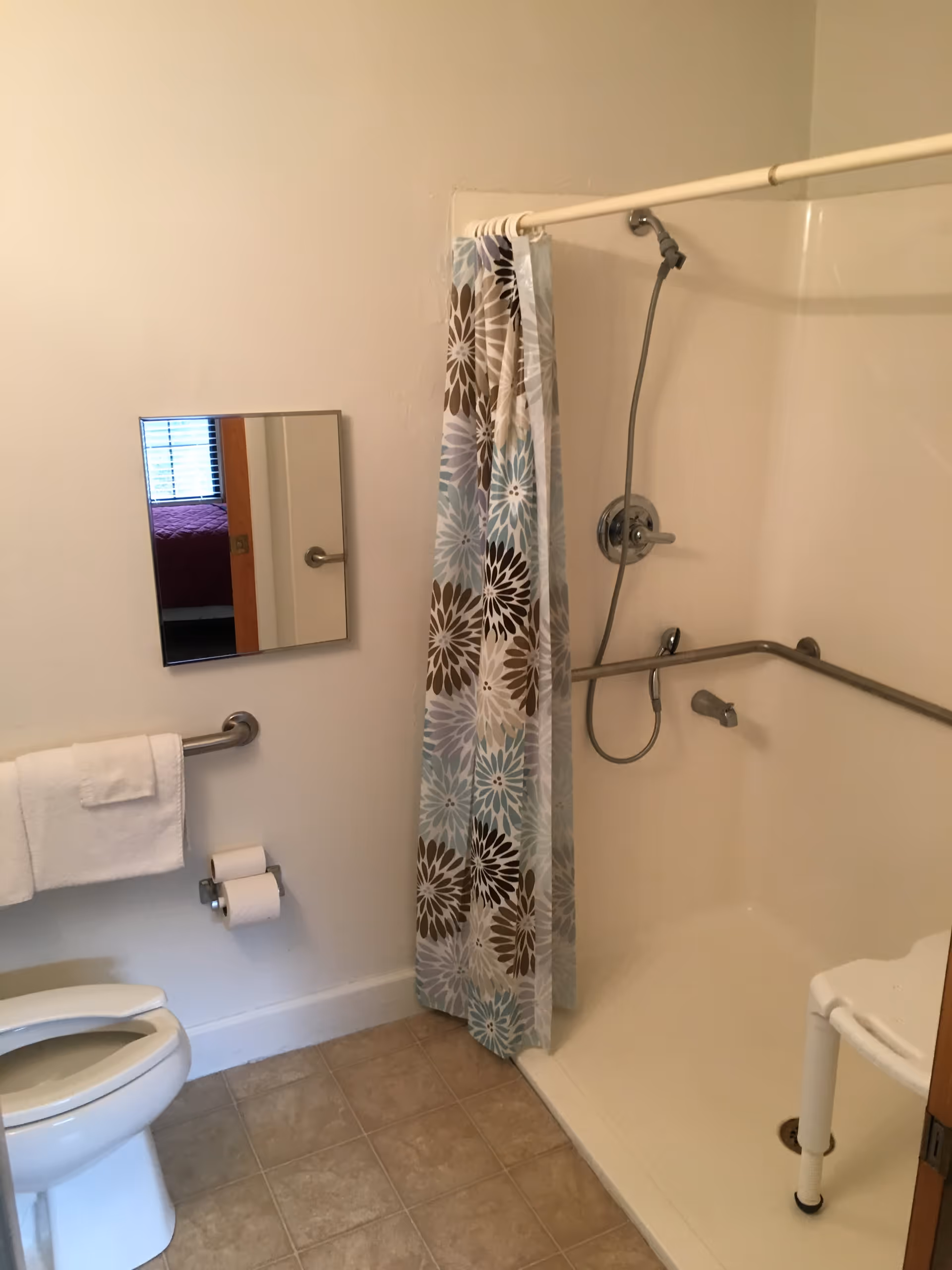 Accessible bathroom with a shower stall, floral shower curtain, grab bars, a shower chair and a toilet.