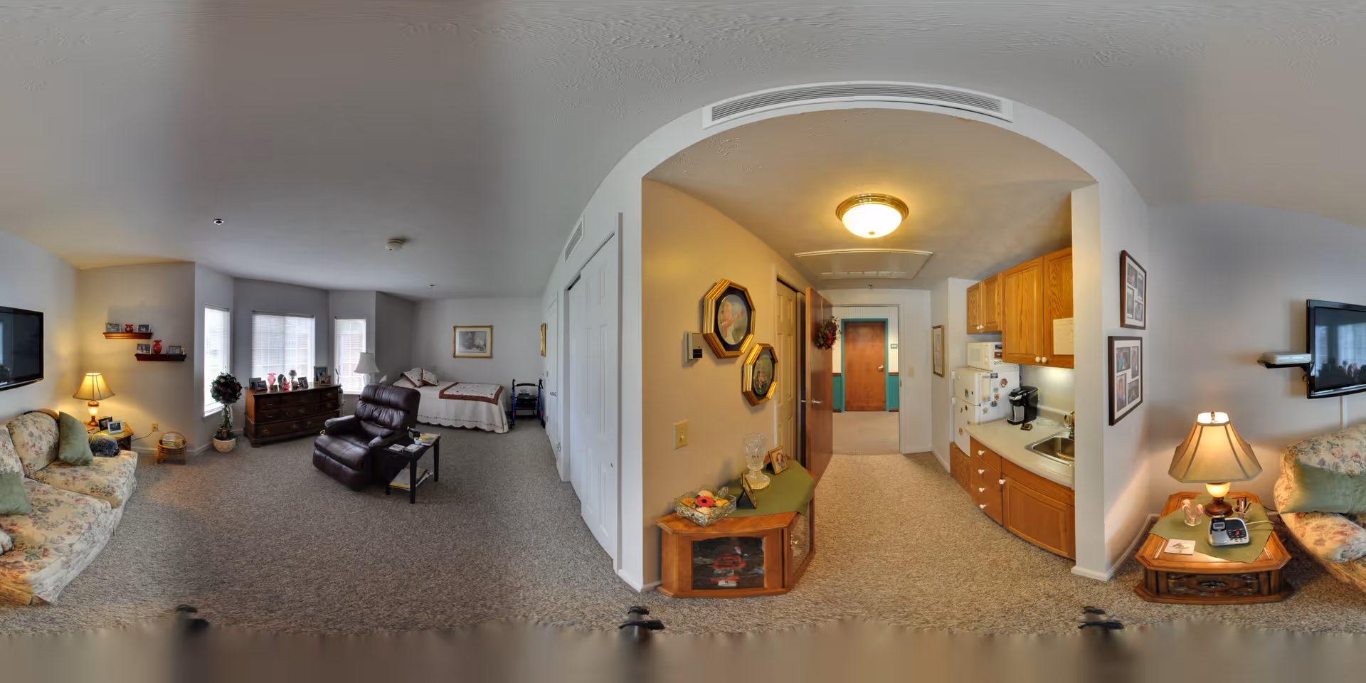 Panoramic interior of a senior living studio showing a living area with sofas and a recliner, a bed in the background, and a kitchenette with an entry hallway.