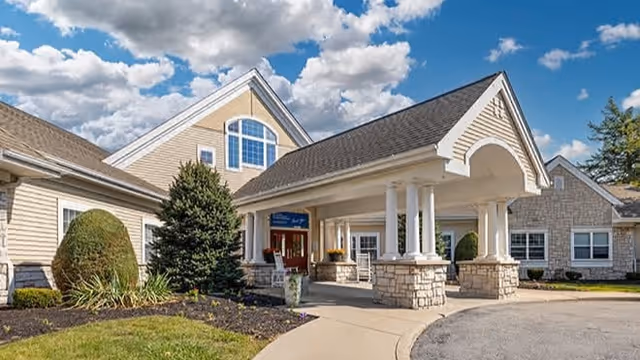 Exterior view of Brookdale Williamsville senior living facility showing a covered entrance with white columns, stone pillars, beige siding, and a well-maintained garden under a partly cloudy sky.