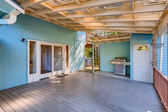 Covered outdoor patio area with wooden flooring and ceiling beams, light blue walls, a stainless steel barbecue grill, a white door, and glass double doors leading inside.