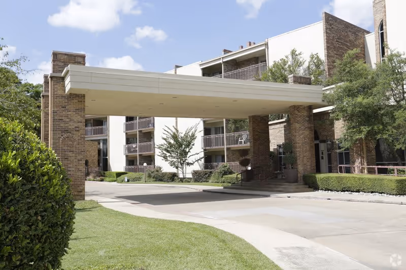 Exterior view of a senior living facility named Three Fountains showing a covered entrance with brick pillars, balconies, trees, and well-maintained landscaping under a partly cloudy sky.