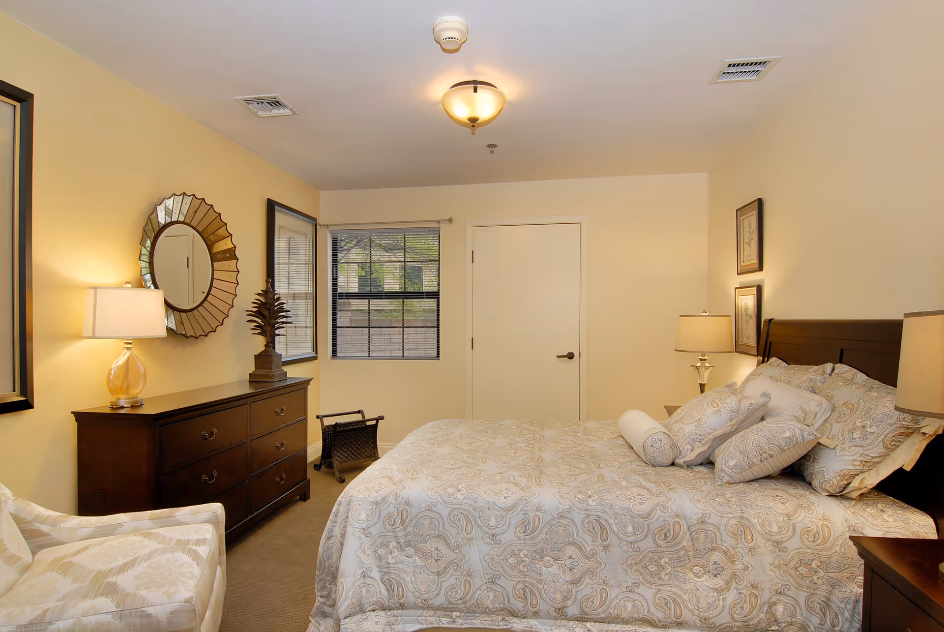 A cozy bedroom with a large bed covered in patterned bedding and multiple pillows. There are two bedside tables with lamps, a wooden dresser with a decorative mirror above it, a window with blinds, and a cushioned armchair in the corner. The walls are painted a soft yellow, and the room has a warm, inviting atmosphere.