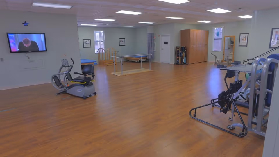 A spacious exercise room with wooden flooring featuring various fitness equipment including a recumbent exercise bike, parallel bars for walking support, and strength training machines. The room has light-colored walls with framed pictures, a wall-mounted TV, and large windows allowing natural light to enter.