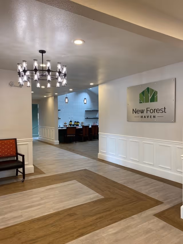 Interior view of a senior living facility hallway with a decorative chandelier, a chair with red upholstery, and a sign on the wall that reads 'New Forest Haven'. The hallway leads to a dining area with tables and chairs visible in the background.
