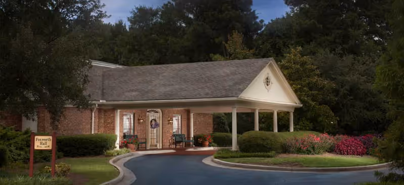 Exterior view of a single-story brick building with a covered entrance supported by white columns. There is a curved driveway leading to the entrance, surrounded by well-maintained bushes and colorful flowers. A sign near the entrance reads 'Foxworth Hall'. Trees and greenery are visible in the background under a partly cloudy sky.