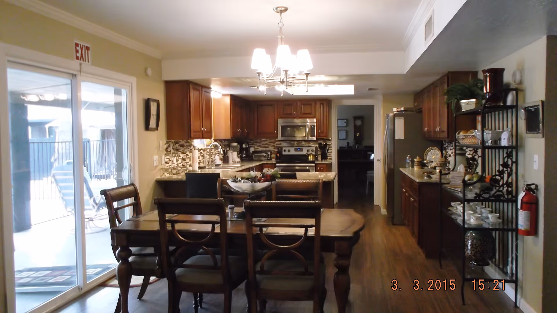 Interior view of a kitchen and dining area with wooden cabinets, stainless steel appliances, a dining table with chairs, a chandelier, and a sliding glass door leading outside.