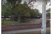 Outdoor view of Bay Gardens Retirement Village showing a grassy area with several trees, a brick wall in the foreground, and a gazebo structure in the middle. The sky is partly cloudy and there is a utility pole on the right side of the image.