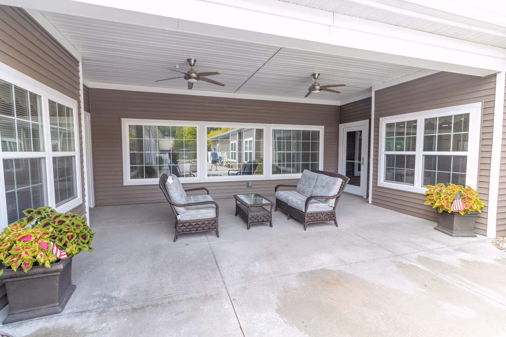 Covered outdoor patio area with two ceiling fans, a cushioned loveseat, two cushioned chairs, and a glass-top coffee table. There are large windows and a door leading inside, with two planters containing green and red foliage and small American flags on either side.