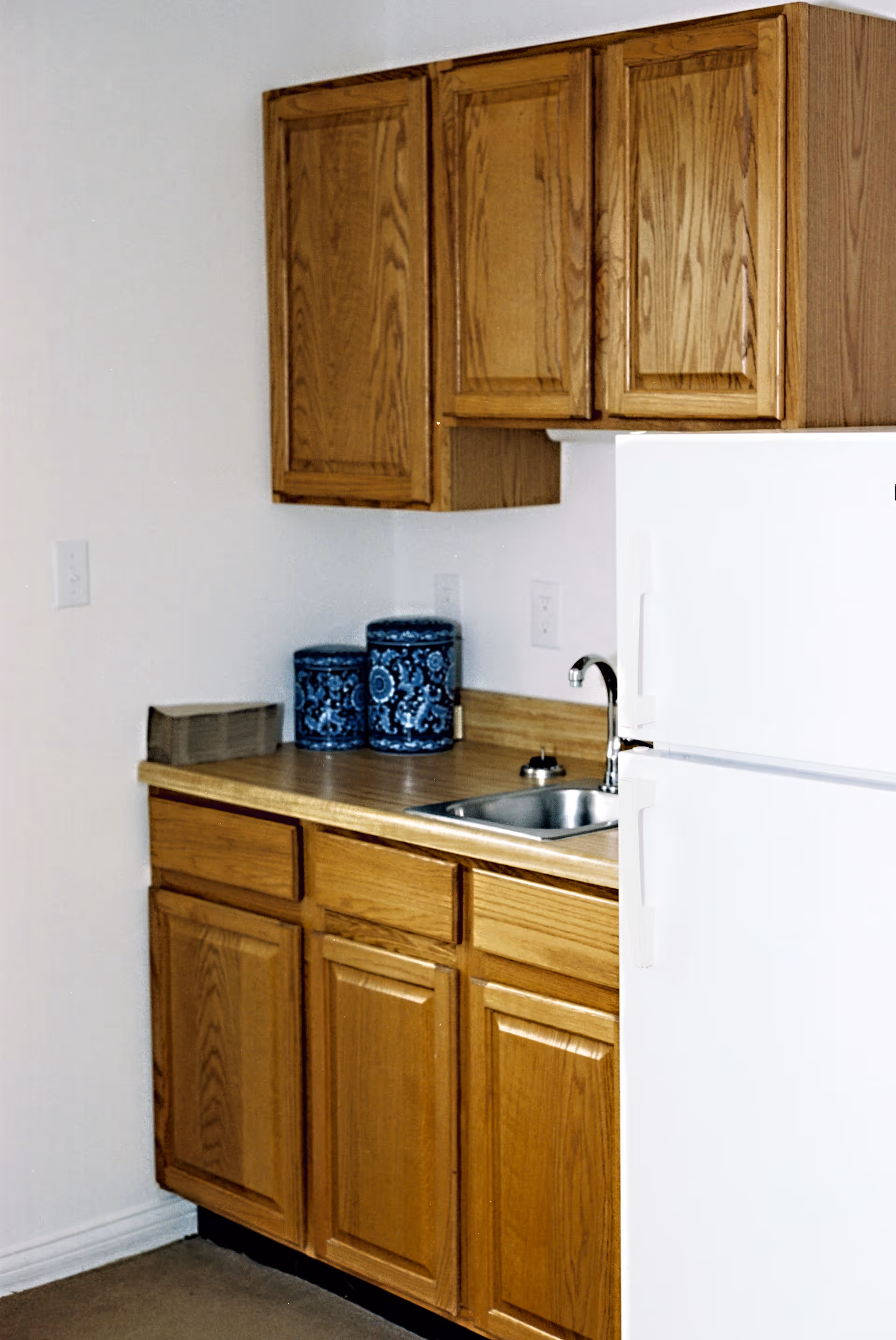 A small kitchen area with wooden cabinets above and below a countertop. On the countertop are two blue decorative canisters and a small sink with a faucet. To the right is a white refrigerator.