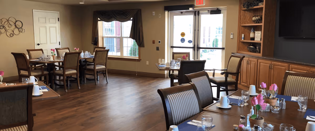 A dining room in a senior living facility with several wooden tables and chairs arranged neatly. Each table is set with placemats, cups, glasses, and small flower decorations. The room has wooden flooring, a large window with curtains, and a glass door leading outside. There is a built-in wooden cabinet with shelves on the right side of the room.