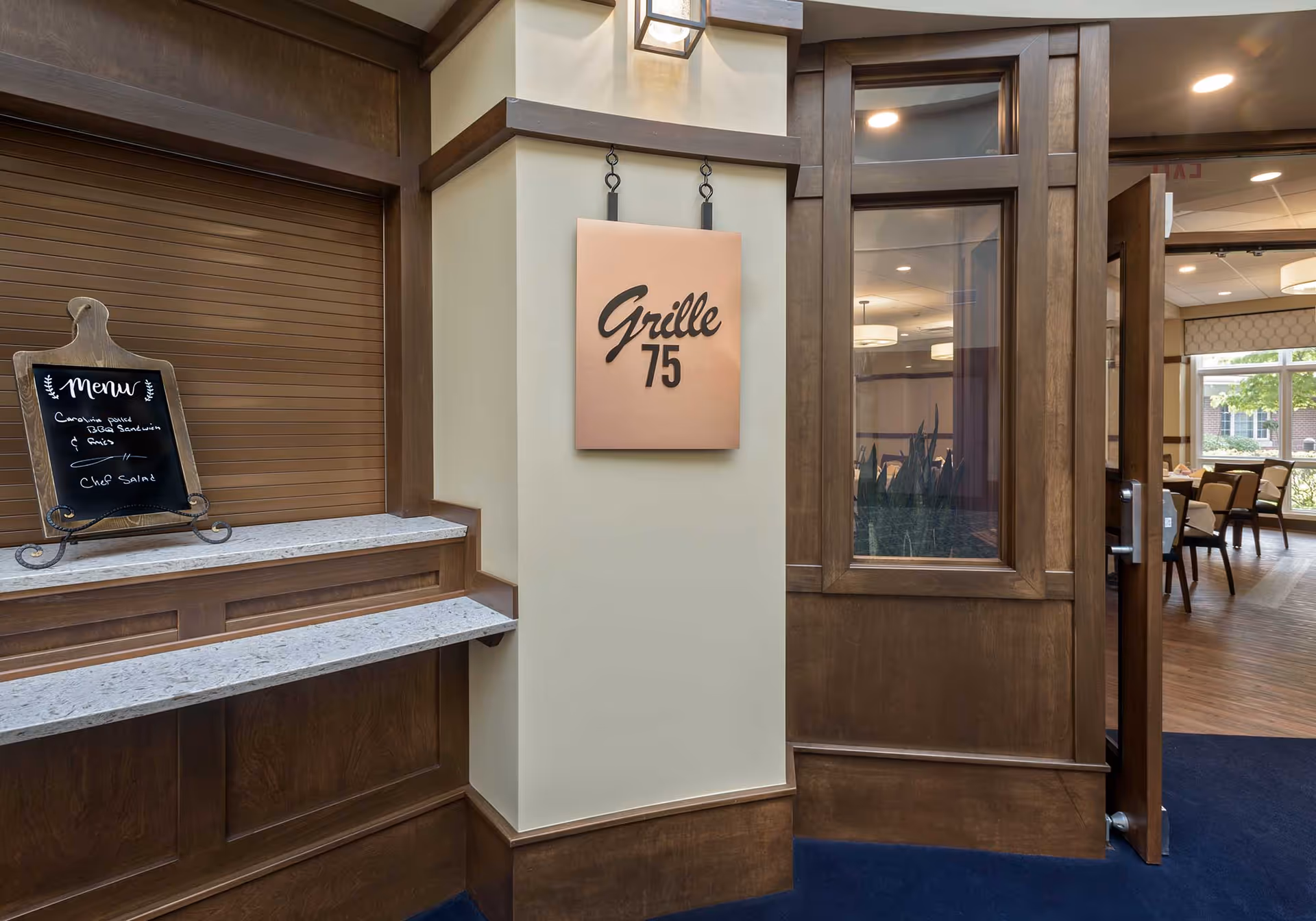 Entrance to a dining area named Grille 75 with a wooden counter and a small chalkboard menu listing Caesar salad, BBQ sandwich & fries, and chop salad. The door to the dining room is open, showing tables and chairs inside with large windows letting in natural light.