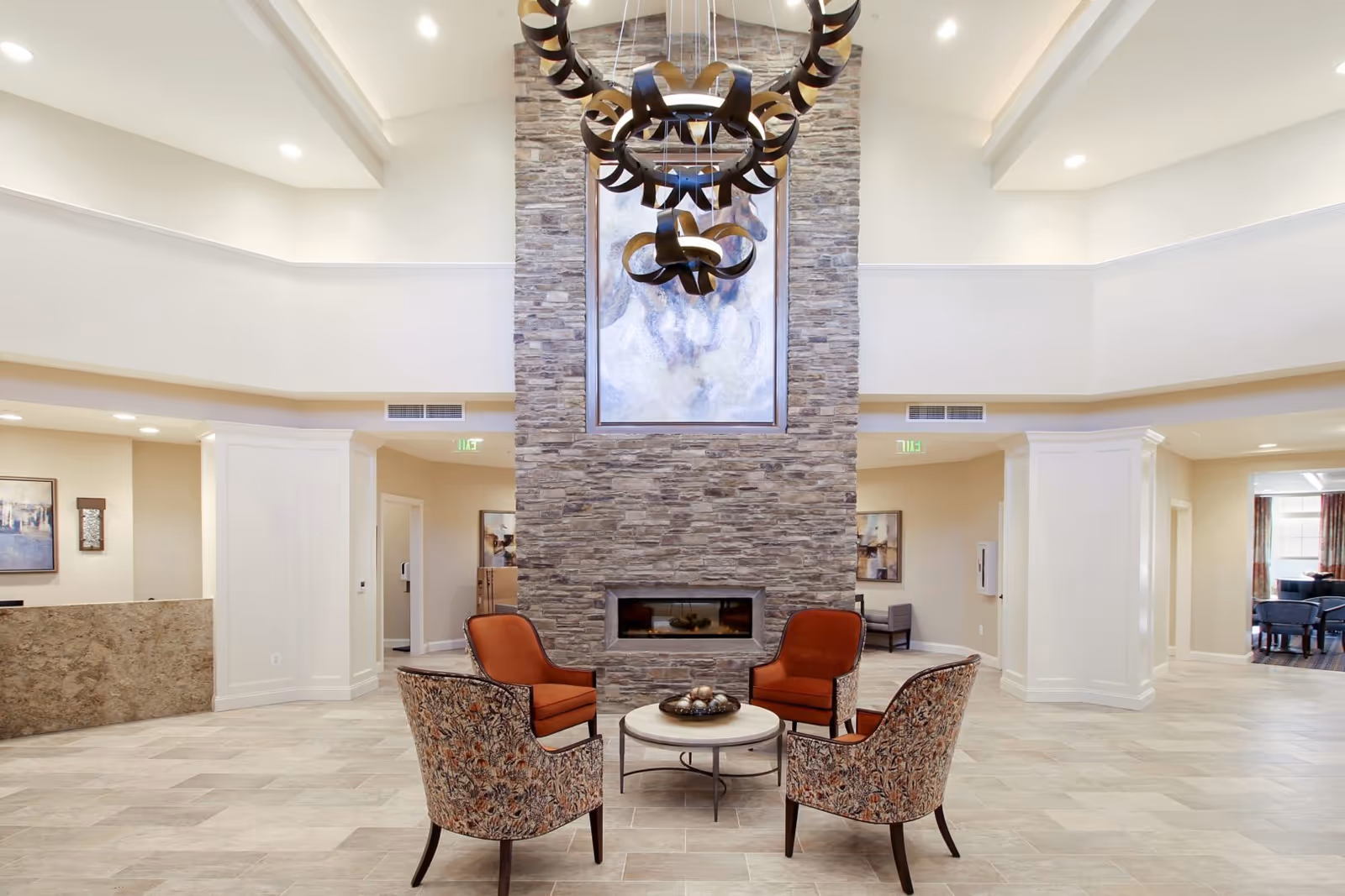 A spacious senior living facility common area with a high ceiling and a large stone fireplace in the center. Four chairs with patterned upholstery and orange cushions are arranged around a small round table with decorative items. Above the fireplace hangs a large painting of a horse, and a modern chandelier with circular elements is suspended from the ceiling. The room features light-colored tiled flooring and neutral walls with recessed lighting.