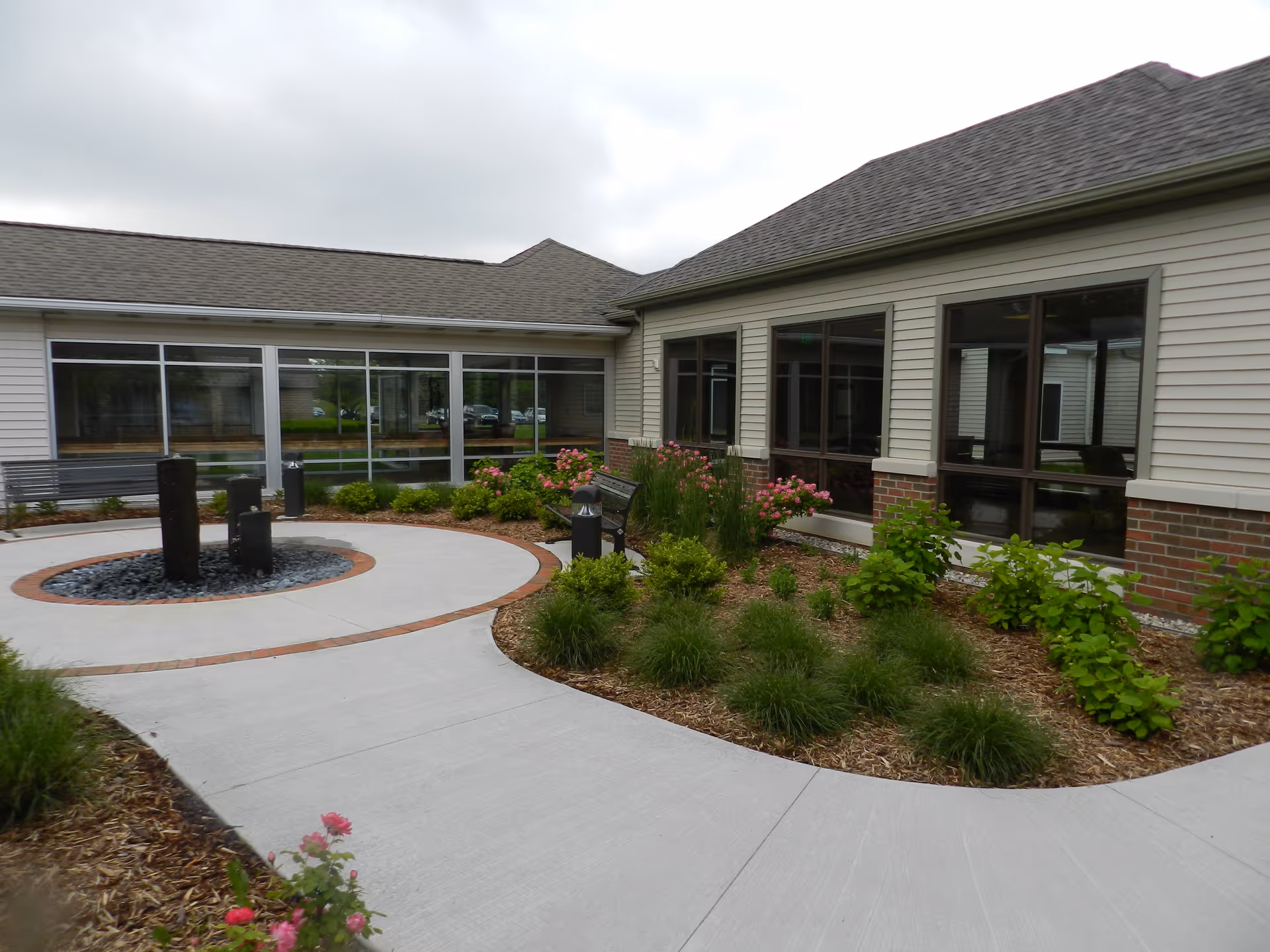 Outdoor courtyard area of a senior living facility with a circular concrete pathway surrounding a small water fountain feature. The building has large windows and beige siding with brick accents. There are landscaped garden beds with green shrubs and pink flowers along the pathway.