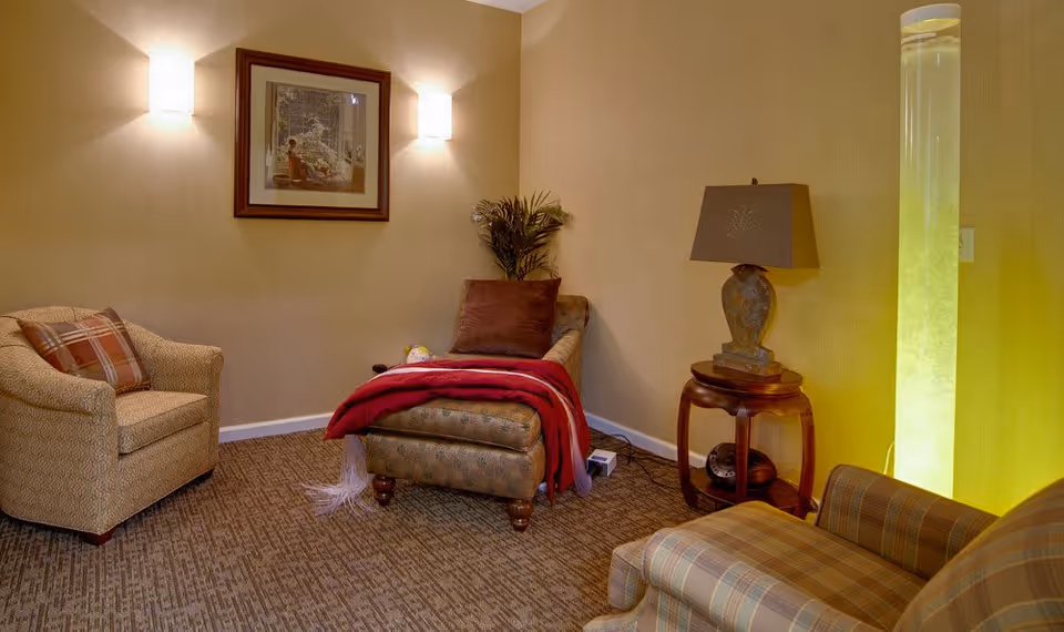 A cozy living room area with a patterned armchair on the left, a cushioned chaise lounge in the center draped with a red blanket and a brown pillow, and a plaid armchair on the right. A small wooden side table with a decorative lamp sits next to the chaise lounge. The walls are beige with two wall sconces illuminating a framed picture. A tall cylindrical light fixture emits a soft yellow glow in the corner.