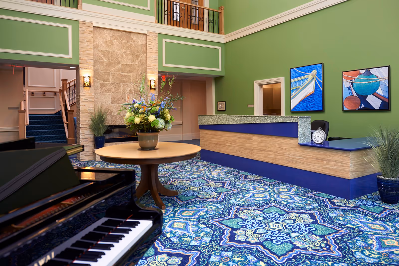 Interior view of a senior living facility lobby with green walls and a patterned blue carpet. A wooden reception desk with a blue countertop is on the right, decorated with a clock and two framed nautical-themed paintings above it. In the center, there is a round wooden table with a large floral arrangement. A black piano is partially visible in the foreground on the left. The back wall features a stone accent with two wall sconces and an open doorway leading to a staircase.