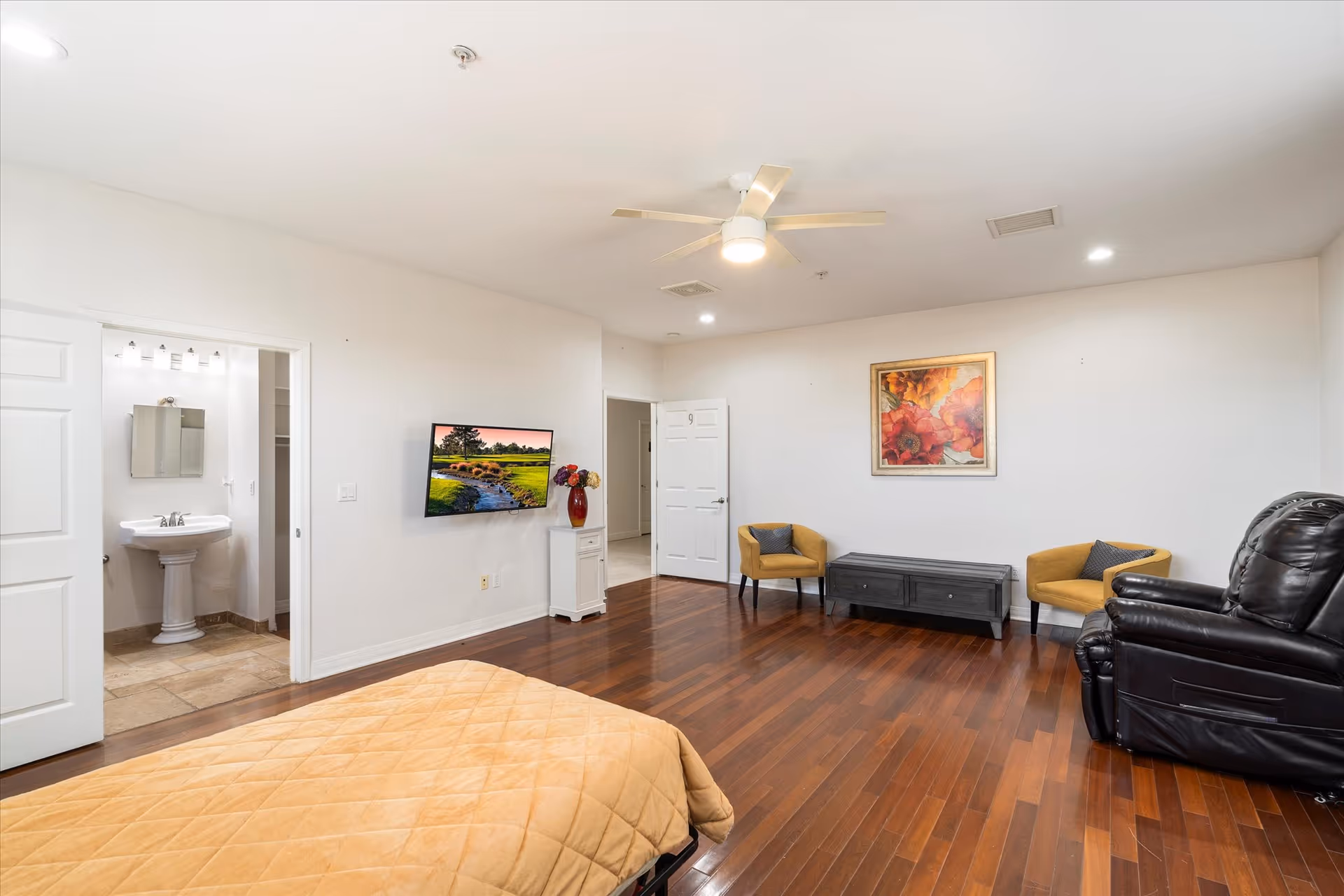 A spacious bedroom with wooden flooring, a bed with a beige quilt, a black recliner chair, two yellow armchairs, a black bench, and a wall-mounted TV displaying a nature scene. There is a ceiling fan with a light, a framed floral painting on the wall, and an open door leading to a bathroom with a pedestal sink and mirror.
