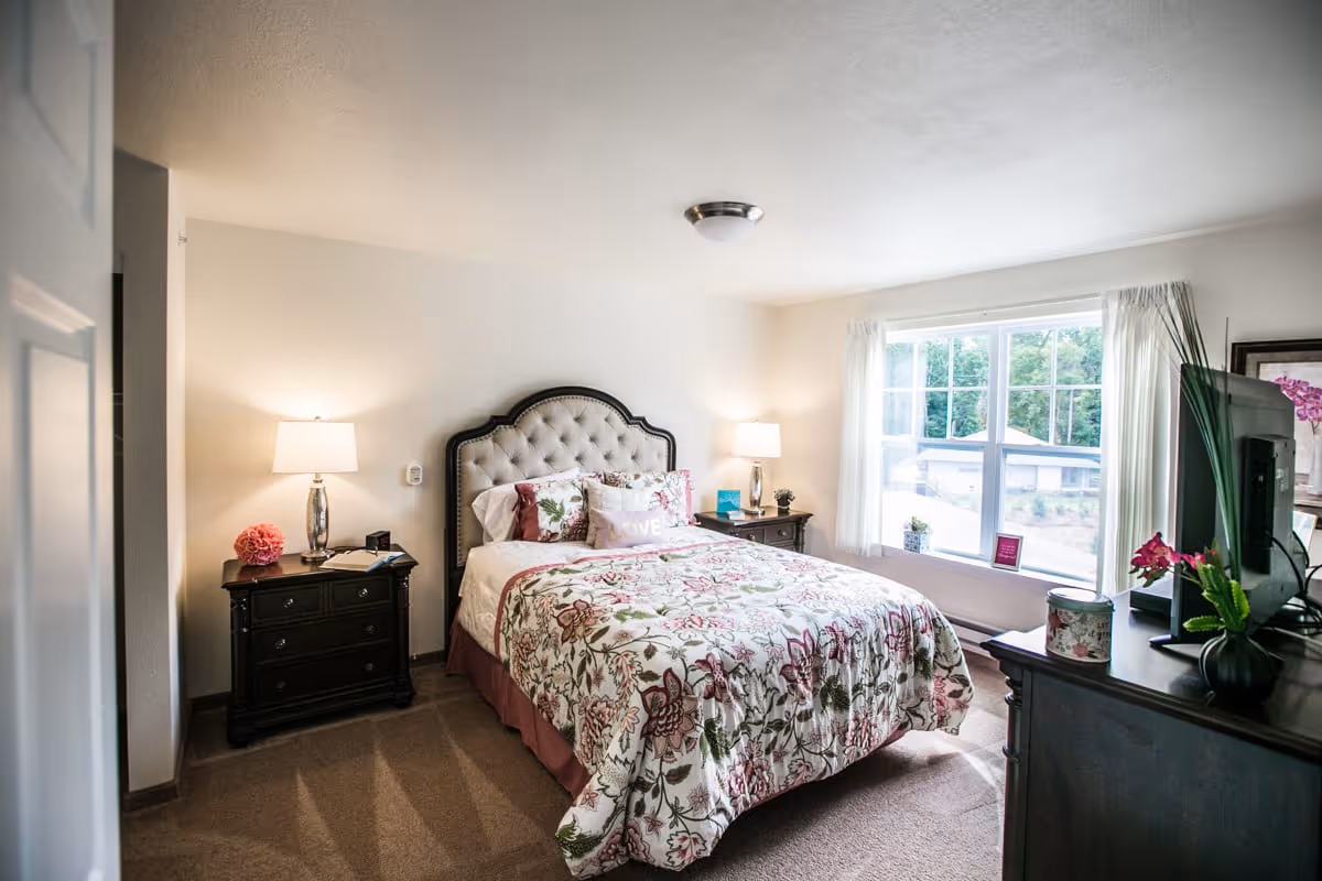 A bright and cozy bedroom with a large window letting in natural light. The room features a bed with a floral patterned comforter and multiple pillows, two bedside tables each with a lamp, and a dresser with a TV and decorative plants. The walls are painted white and the carpet is beige.