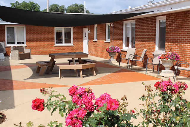 Outdoor courtyard area with a picnic table and benches under a large black shade sail. The courtyard is surrounded by a red brick building with several windows and a white door. There are flower pots with pink flowers placed near the building walls and vibrant pink flowers in the foreground.