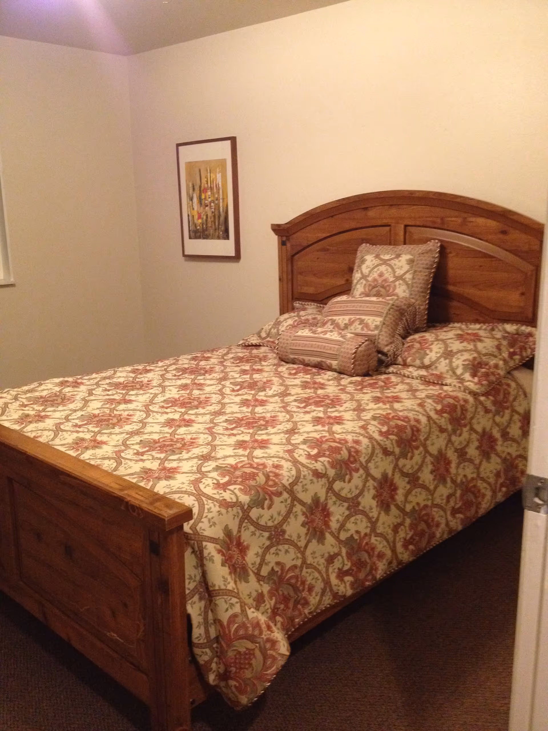 A bedroom with a wooden bed frame and a bed covered with a floral patterned bedspread in shades of red and beige. There are multiple matching pillows on the bed. A framed abstract painting hangs on the beige wall to the left of the bed.