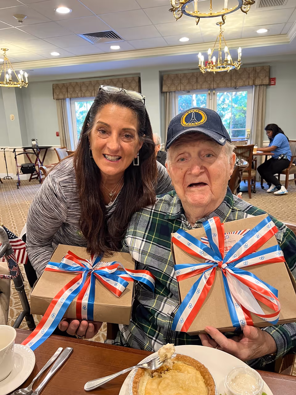An elderly man and a woman in a dining room holding wrapped gift boxes tied with red, white, and blue ribbons.