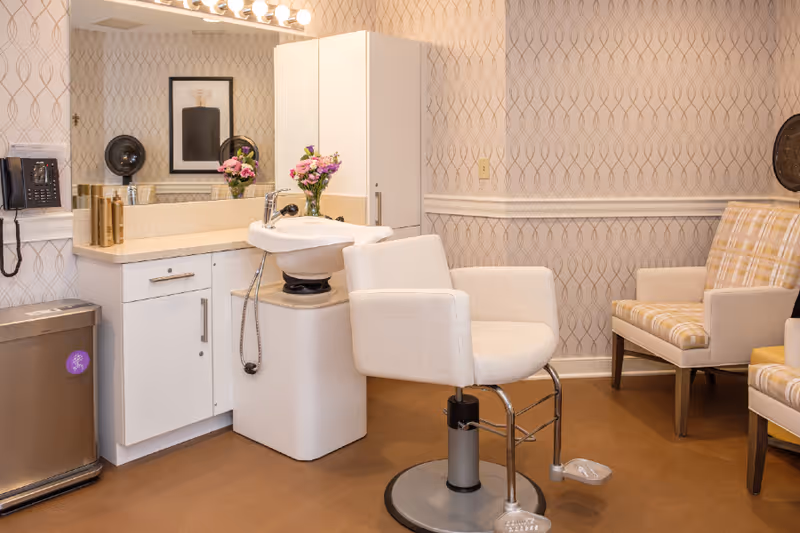 A cozy salon area in a senior living facility featuring a white salon chair in front of a white sink with a faucet. The room has patterned wallpaper, a large mirror with lights above it, a white cabinet, a wall-mounted black telephone, a trash bin, and two cushioned chairs with plaid upholstery. A vase with pink flowers is placed on the countertop.