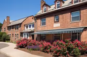 Exterior view of a brick senior living facility building with multiple windows and a green and white striped awning. There are vibrant pink flowers and greenery planted along the walkway in front of the building under a clear blue sky.