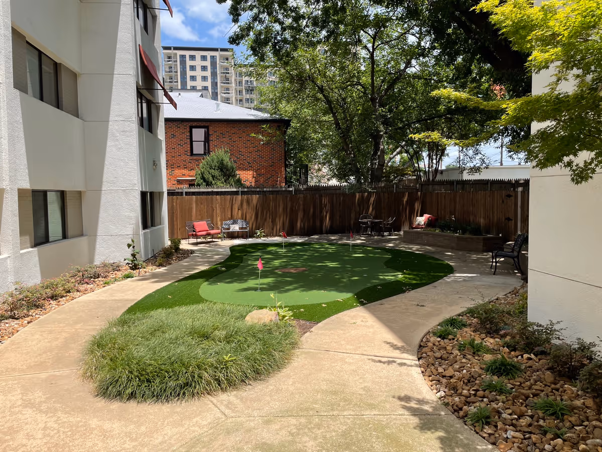 Outdoor courtyard area with a small artificial putting green surrounded by a curved concrete walkway. There are benches and chairs with cushions along the fence and near the building walls. Trees and plants provide shade and greenery, with a brick building and a taller apartment building visible in the background under a partly cloudy sky.