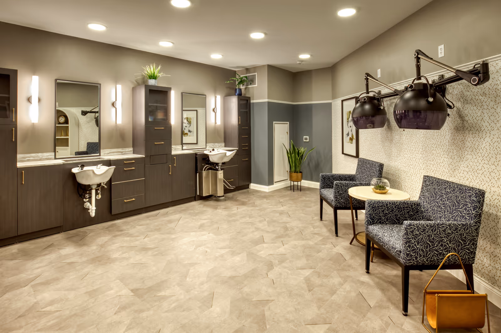 A modern salon area in a senior living facility with two hair washing sinks, large mirrors, dark wood cabinetry, and two black hair dryers mounted on the wall above two patterned armchairs and a small round table with a decorative glass bowl. The room has beige and gray walls, recessed ceiling lights, and a potted plant in the corner.