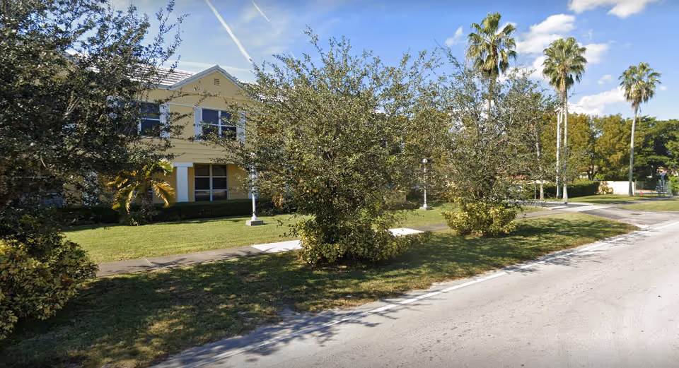 Exterior view of a yellow two-story building partially obscured by trees and bushes, with a sidewalk and a road in the foreground under a blue sky with some clouds.