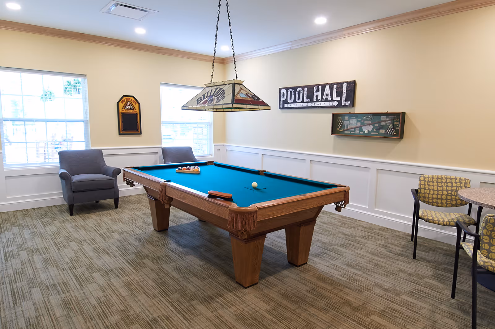 A well-lit room with a pool table in the center, two gray armchairs near two large windows, a hanging light fixture above the pool table, and a sign on the wall that reads 'POOL HALL Rack It & Crack It'. There is also a small round table with two patterned chairs on the right side of the room.