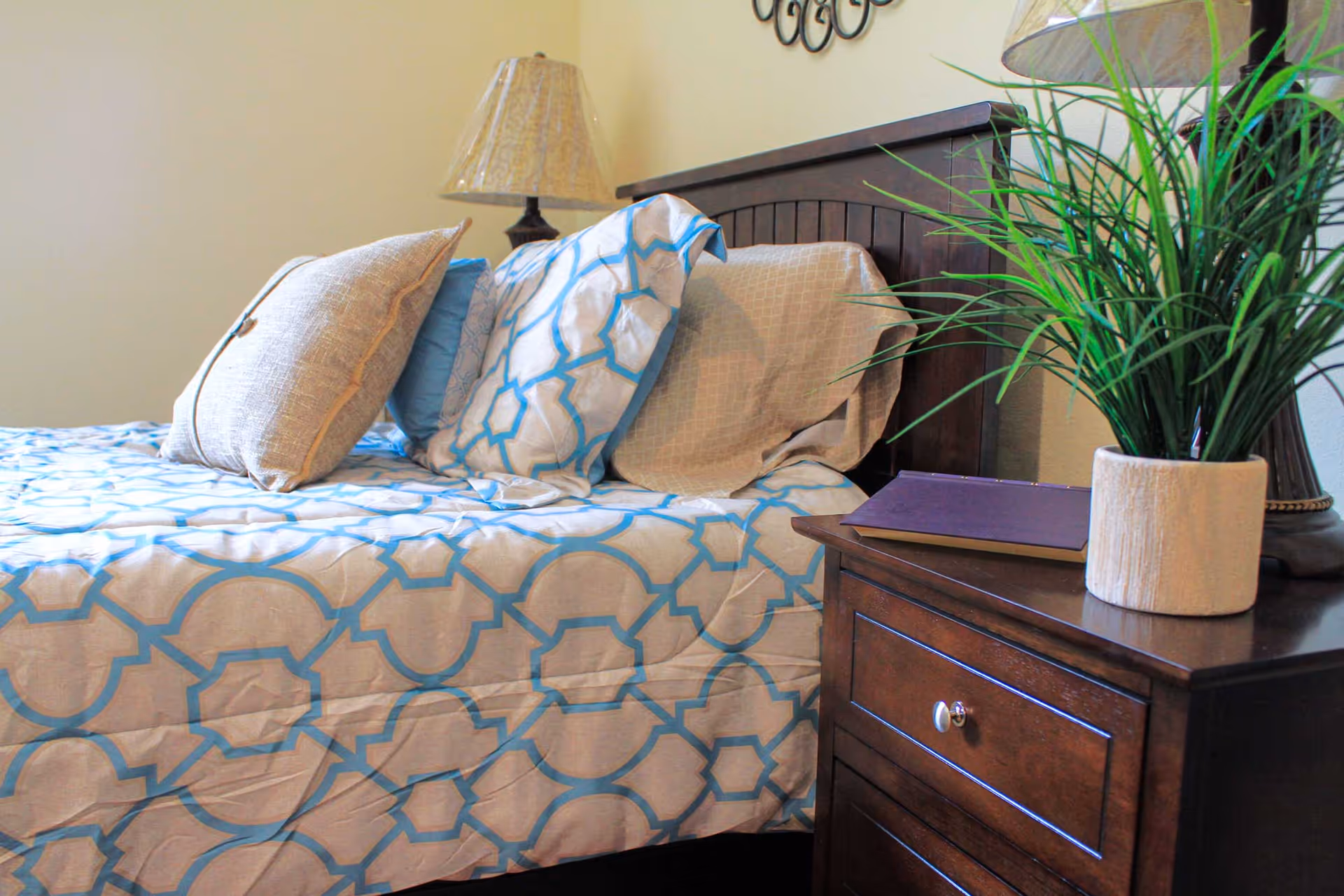 A neatly made bed with a blue and beige patterned bedspread and several pillows, next to a wooden nightstand with a potted plant, a closed book, and a lamp with a beige lampshade.