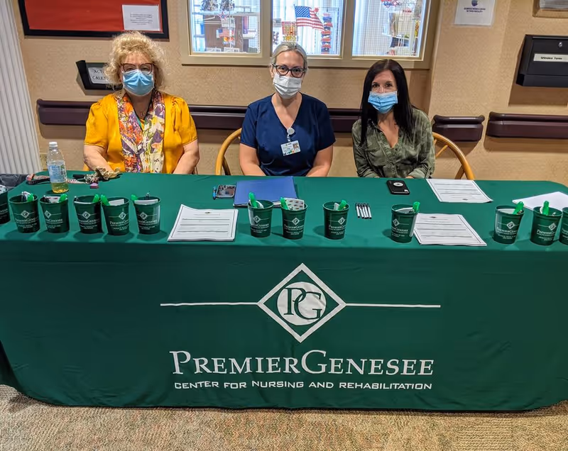 Three women wearing face masks sitting behind a table covered with a green tablecloth that reads 'Premier Genesee Center for Nursing and Rehabilitation.' The table has several green cups with pens and papers on it. Behind them is a beige wall with windows and some posted notices.