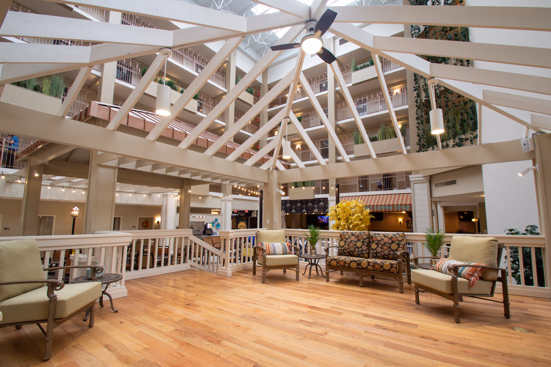 Spacious indoor common area with wooden flooring, cushioned chairs, and a patterned sofa arranged around small tables. The area is covered by a large glass ceiling with white beams, allowing natural light to illuminate the space. Multiple floors with balconies and plants are visible in the background.
