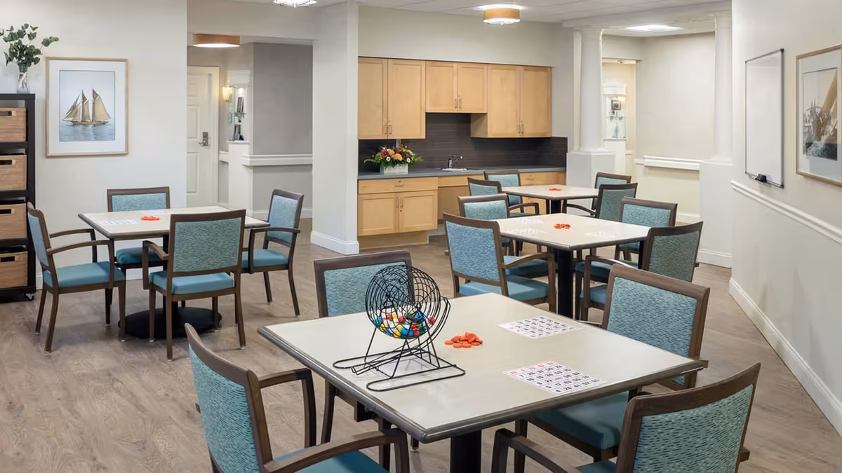 A bright room with multiple tables and chairs arranged for group activities, featuring a bingo cage with colorful balls and bingo cards on the tables. The room has light wood flooring, light-colored walls, a kitchenette with wooden cabinets, and framed artwork on the walls.