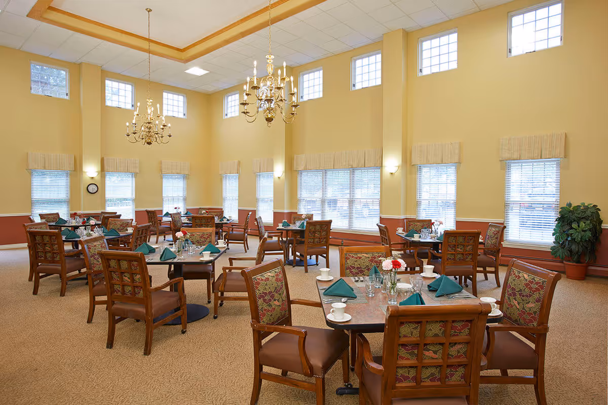 A spacious dining room with multiple wooden tables and chairs arranged neatly. Each table is set with green folded napkins, glassware, white cups, and small flower vases. The room has high ceilings with two ornate chandeliers and several windows allowing natural light to fill the space. The walls are painted yellow with a reddish-brown lower section, and there is a potted plant in the corner.