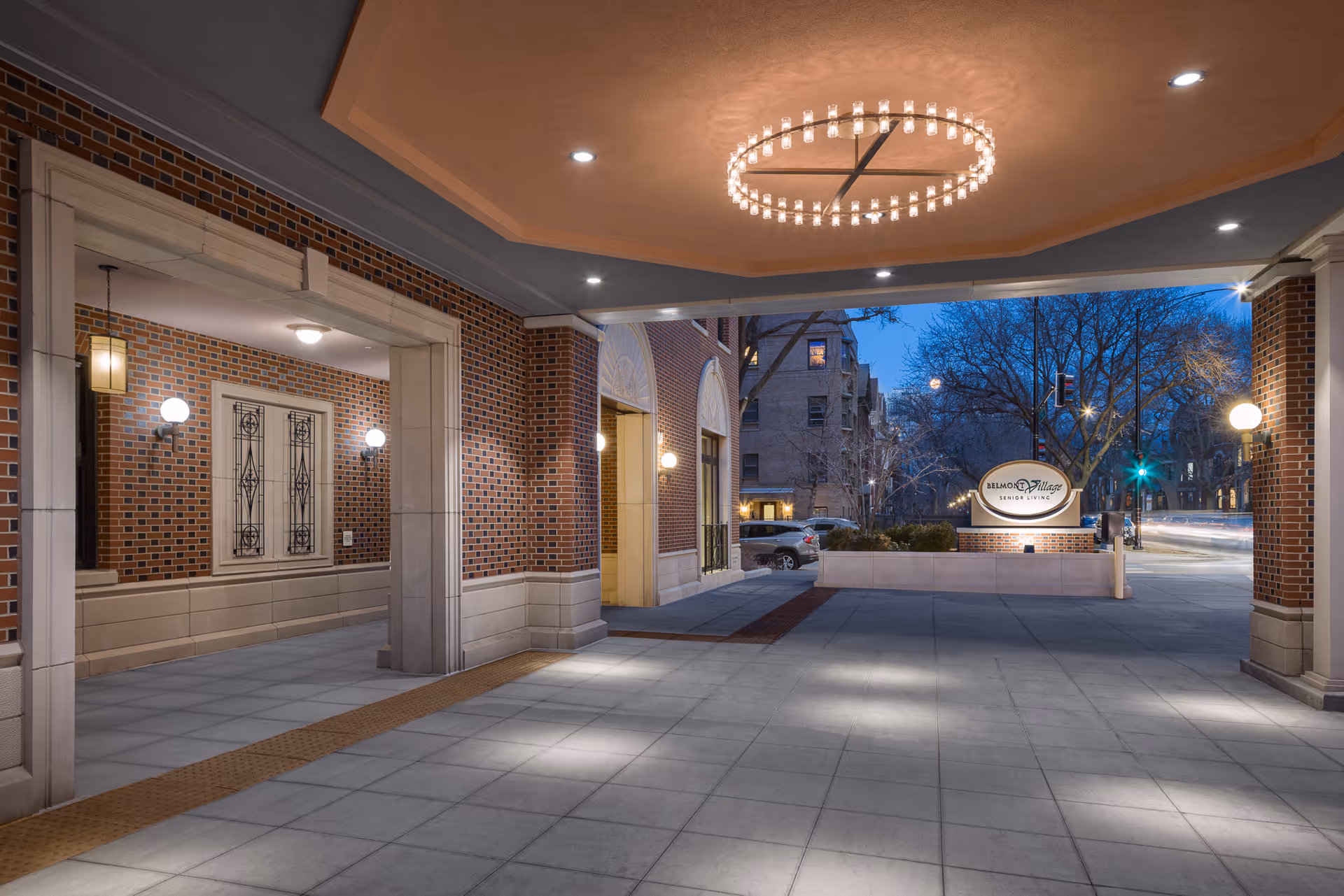 Evening view of the entrance area of Belmont Village Senior Living Lincoln Park, featuring brick walls, decorative lighting fixtures, a circular chandelier on the ceiling, and a sign with the facility's name visible near the street.