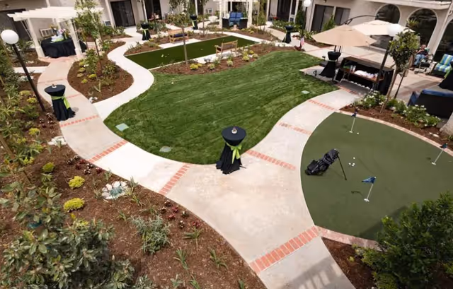 A landscaped courtyard at a senior living facility with winding paved paths, lawns, a small putting green, seating areas and planted beds.