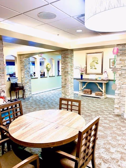 Interior view of a senior living facility common area with a round wooden table and four chairs in the foreground. Stone pillars and archways separate different sections of the room. A console table with decorative items and framed artwork is visible against the far wall. The space is well-lit with ceiling lights and has patterned carpet flooring.