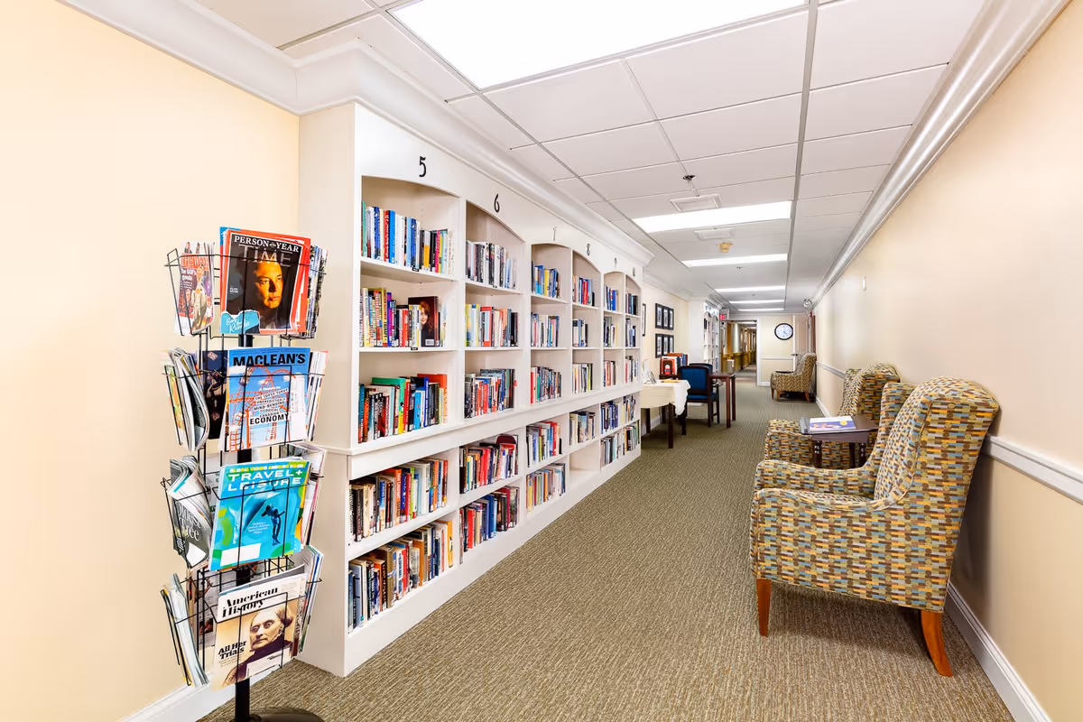 A well-lit hallway in a senior living facility with built-in bookshelves filled with books numbered 5 to 8. On the left side, there is a rotating magazine rack with various magazines. On the right side, there are patterned armchairs with small tables between them. The hallway has beige walls, a carpeted floor, and a ceiling with fluorescent lighting.