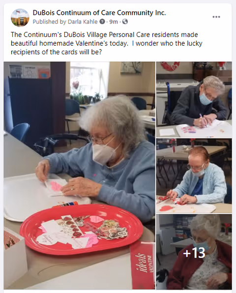 Several elderly individuals wearing masks are seated at tables in a communal room, engaged in making homemade Valentine's Day cards with paper hearts and craft supplies.