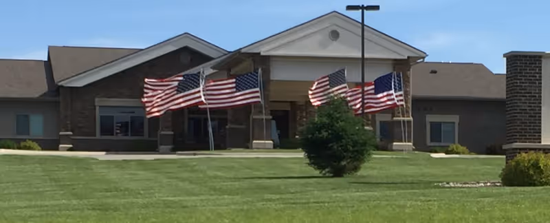 Front entrance of a single-story brick building with several American flags on poles and a manicured lawn.