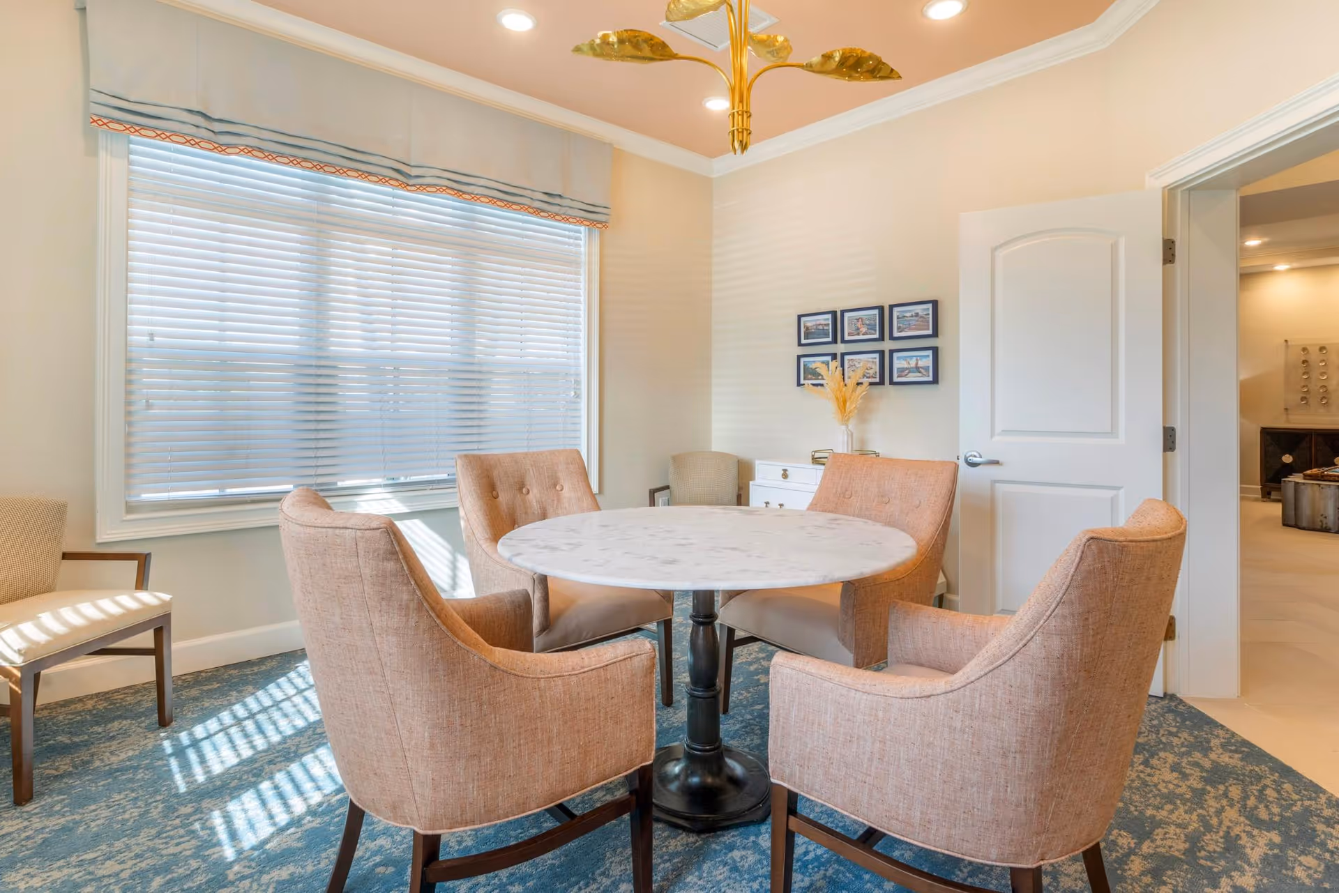 Bright seating area with a round marble table surrounded by four upholstered chairs near a large window with blinds.