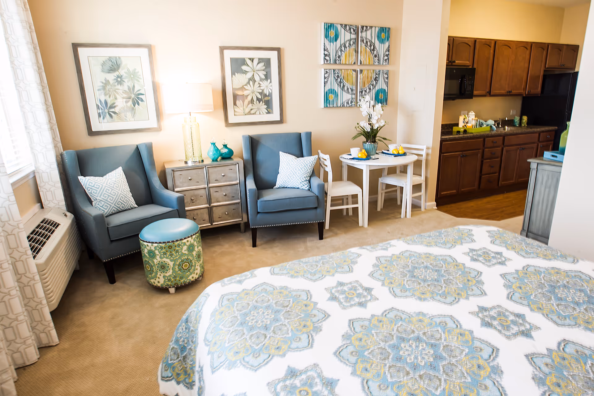 A cozy studio apartment featuring a bed with a patterned blue and yellow bedspread in the foreground, two blue armchairs with patterned pillows, a small round table with two white chairs, decorative wall art, a lamp on a small chest of drawers, and a kitchenette with wooden cabinets and a microwave in the background.