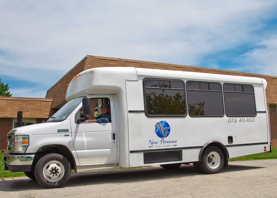 A white shuttle bus parked in front of a brick building under a partly cloudy sky. The bus has the logo and text 'New Florence Care Center' and a phone number (573) 415-9333 on its side. A person is visible sitting in the driver's seat, smiling.